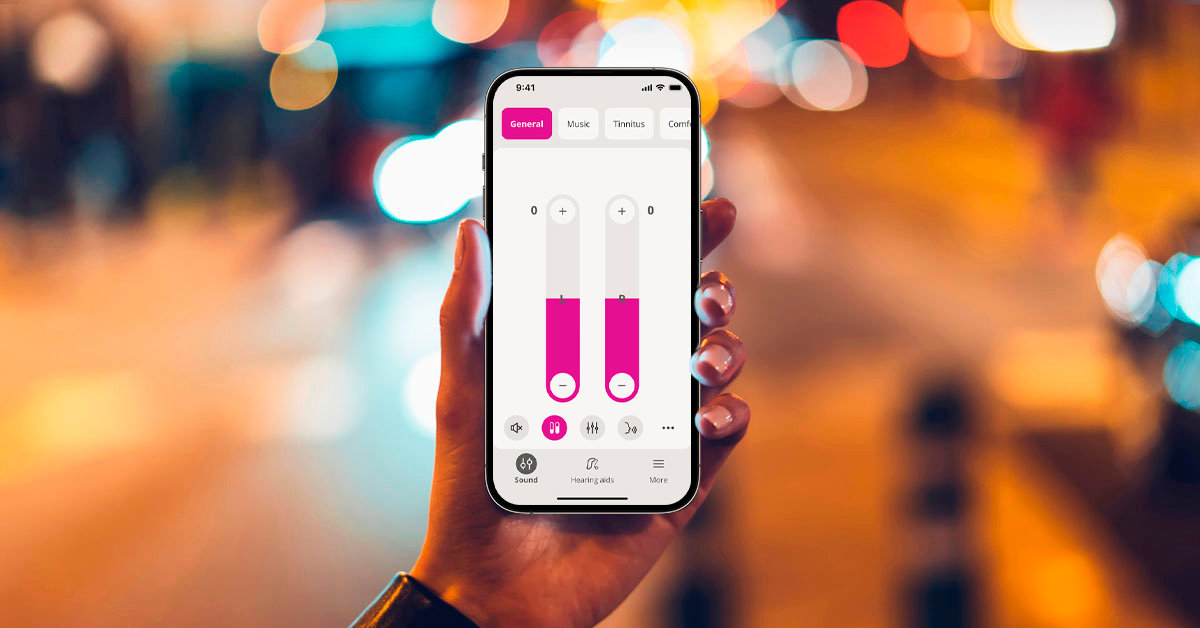The Demant Oticon Companion app showing volume meters on a mobile phone held up by a hand against a colorful background.